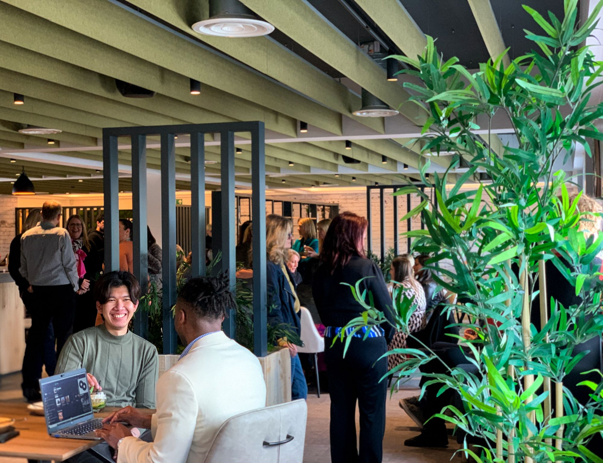 A lively café setting with a mix of people socializing and working. A man in a green shirt smiles at the camera while seated at a table with a laptop. In the background, a group of people engage in conversation, surrounded by greenery and modern decor.
